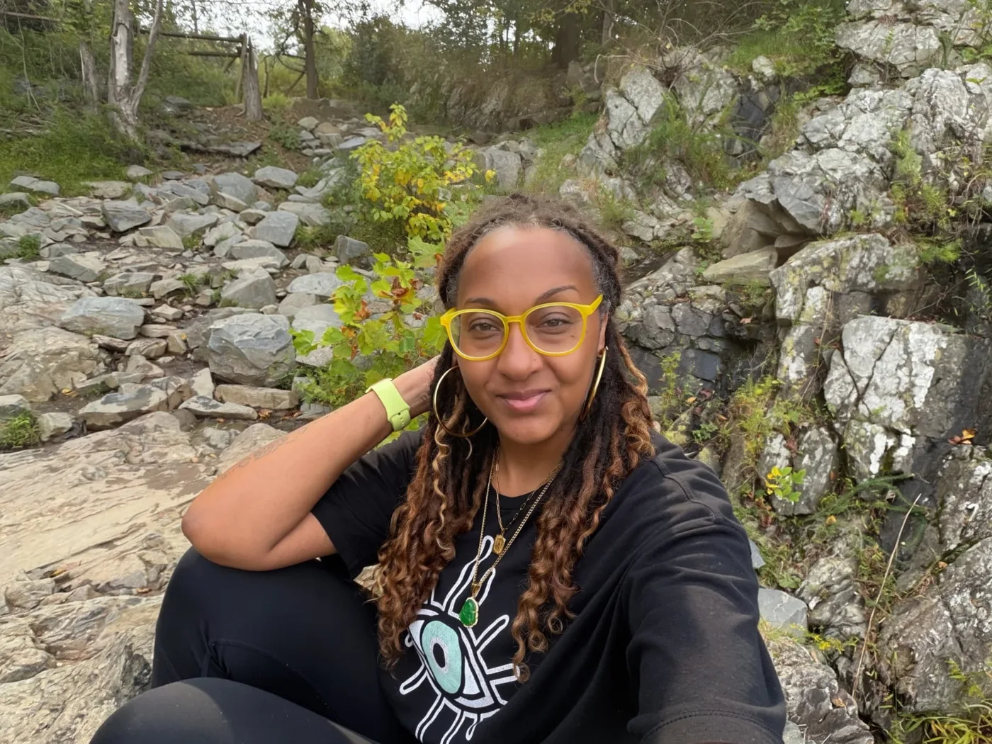Nalani York in yellow glasses sitting outdoors among rocks and greenery.