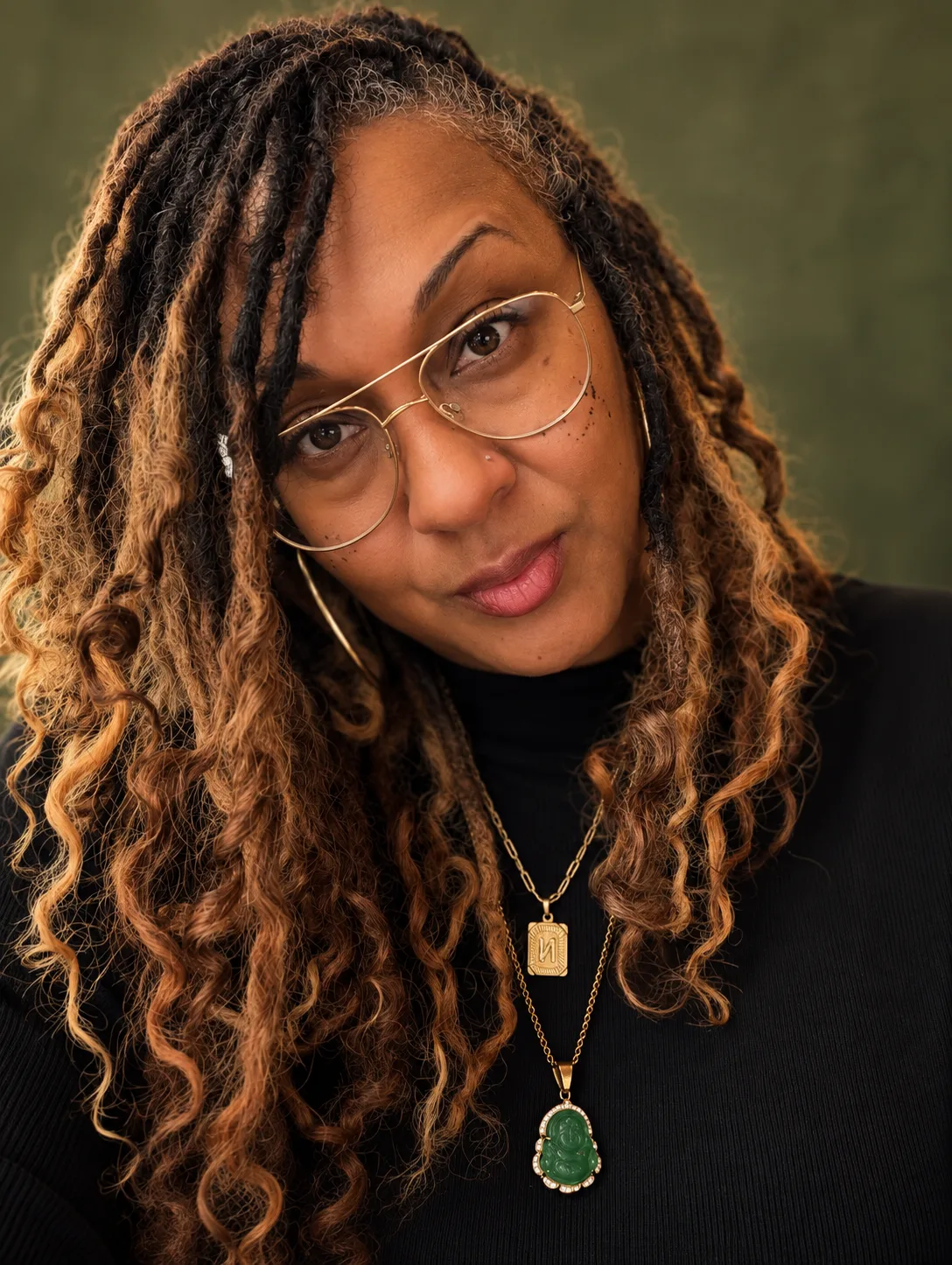 Nalani York in a black turtleneck and gold aviator glasses, wearing a jade Buddha pendant, photographed against a moss green backdrop.
