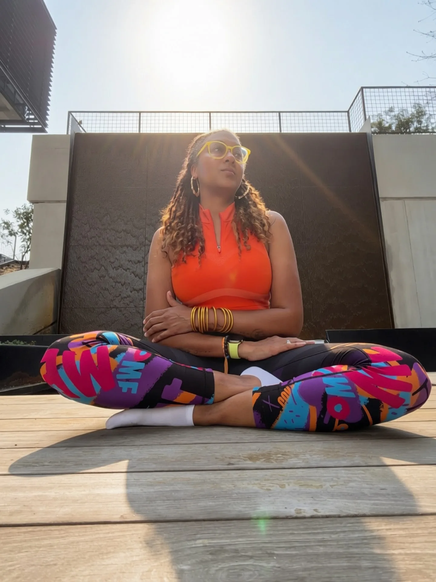 Nalani York seated cross-legged on a wooden deck in front of a water wall, wearing an orange athletic top, printed leggings, yellow glasses and gold hoop earrings, looking upward.